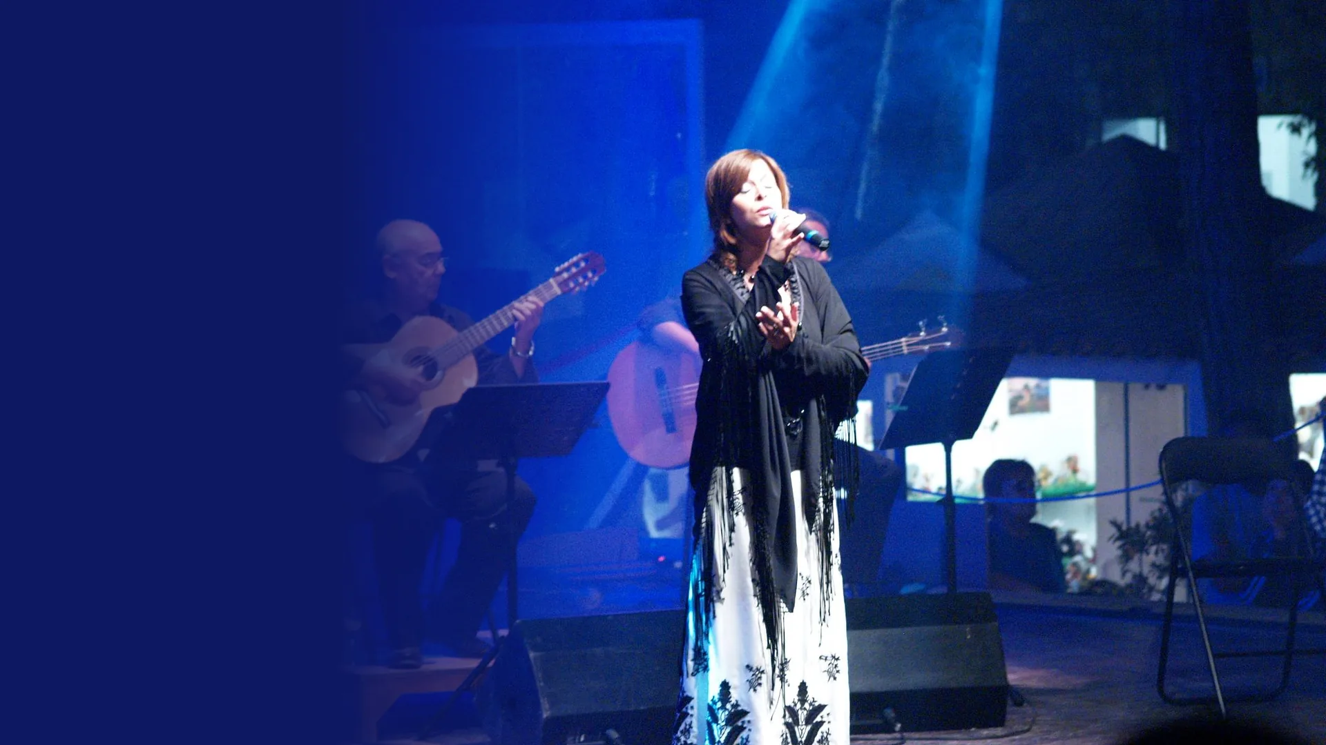 A fadista in a black fringed shawl and long white skirt sings with her eyes closed on a blue-lit stage while two guitarists play behind her.