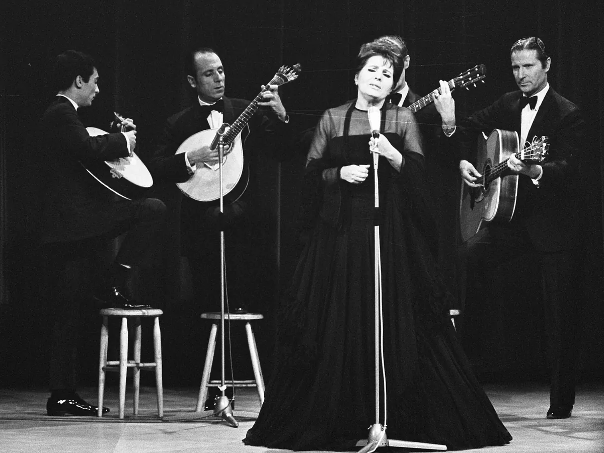 Amália Rodrigues in a long black dress singing at a microphone on a stage, with guitarists in dark suits seated around her.