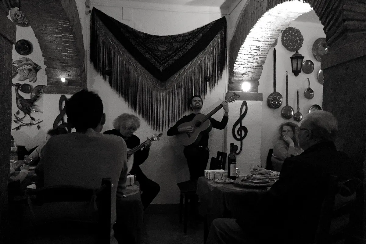 A dim, arched room in Lisbon with a black fringed shawl on the wall and two guitarists playing among diners at small tables.