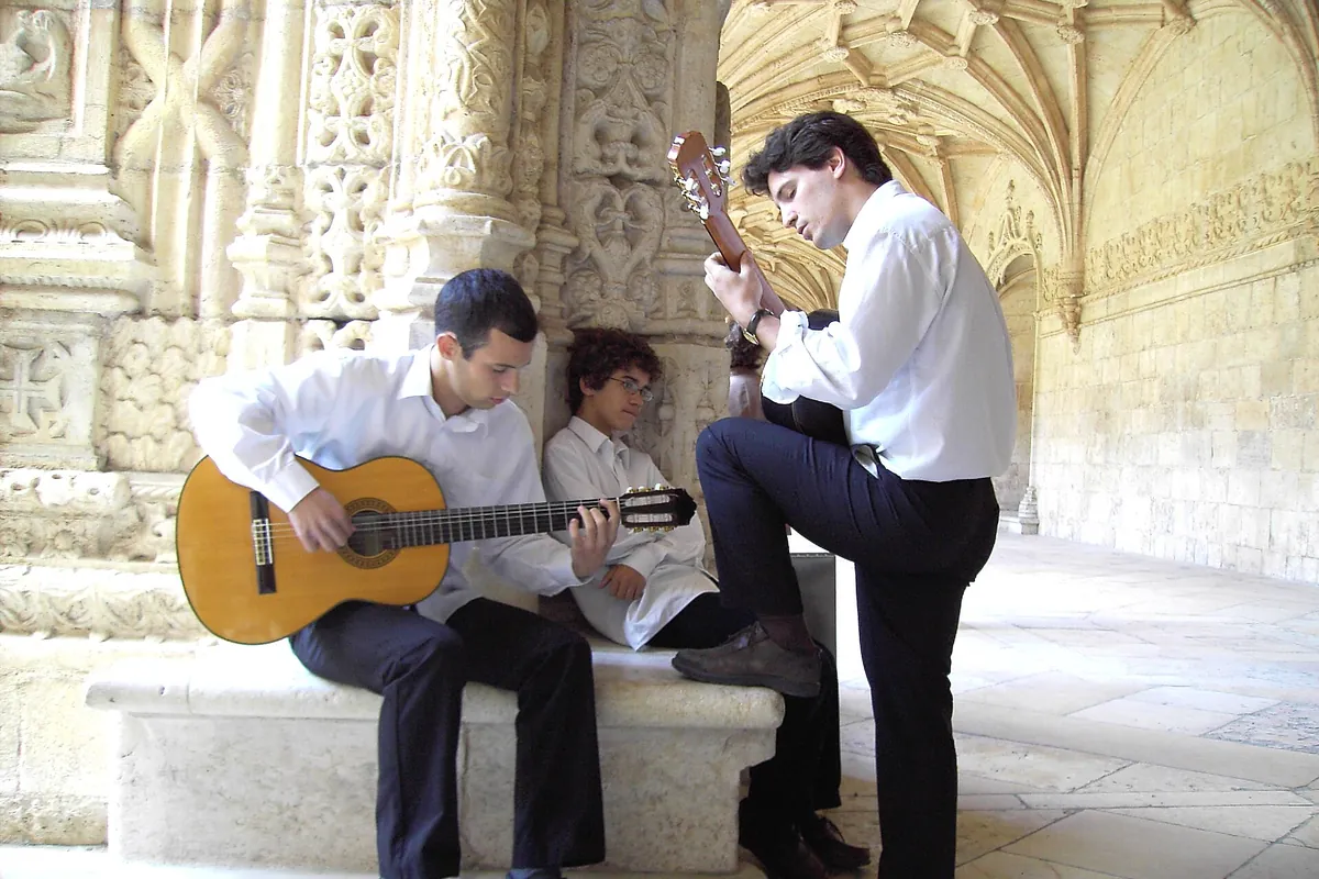 Three young guitarists in white shirts in a stone cloister with carved arches, rehearsing fado.