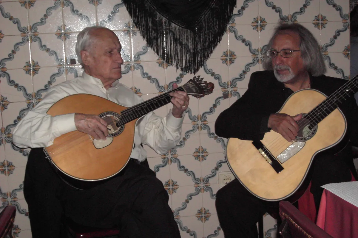 A Portuguese guitar and an acoustic guitar played side by side in front of a tiled wall hung with a black fringed shawl.