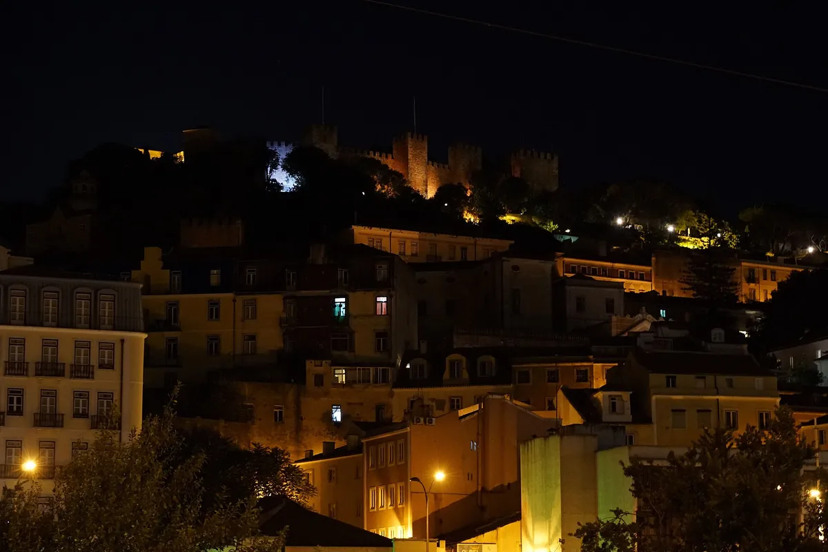 The lit roofs and walls of Lisbon's old quarter at night below a floodlit castle on the hill.